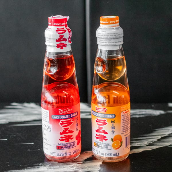 Two Shirakiku Ramune carbonated drinks, strawberry red and orange, on a dark surface.