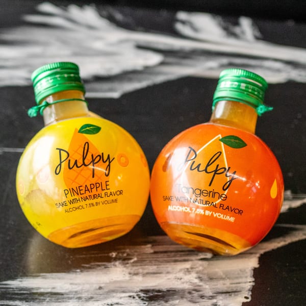 Two small, round bottles of Pulpy sake, pineapple and tangerine flavors, on a dark surface.