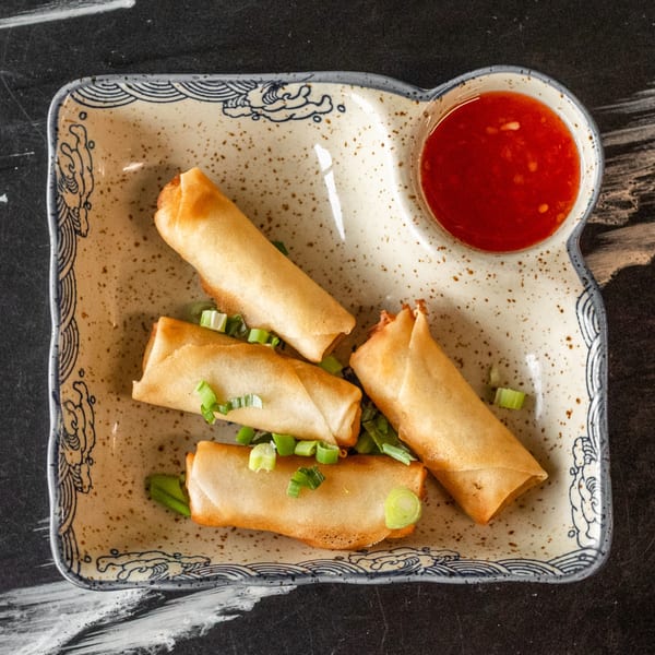 Four crispy spring rolls garnished with green onions, served with a side of red dipping sauce.