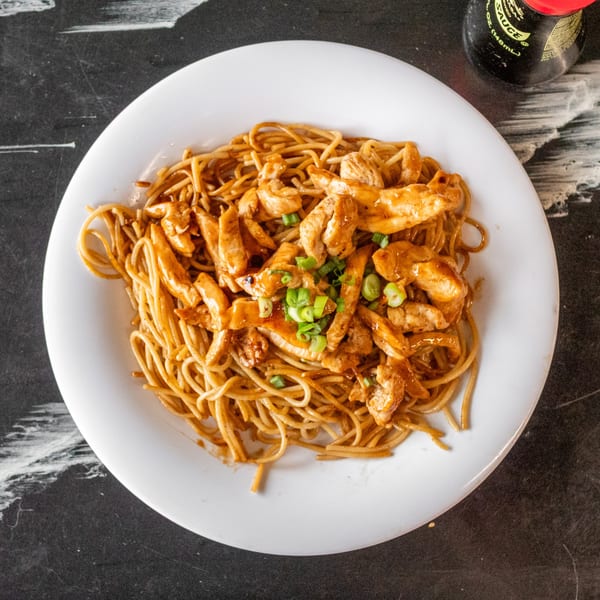 Chicken lo mein noodles with green onions on a white plate, with a soy sauce bottle in the background.