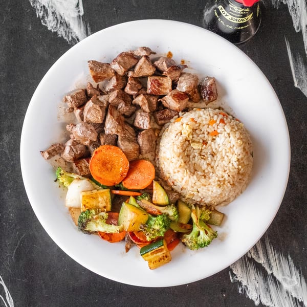 A white plate with steak bites, fried rice, and mixed grilled vegetables, with a soy sauce bottle.