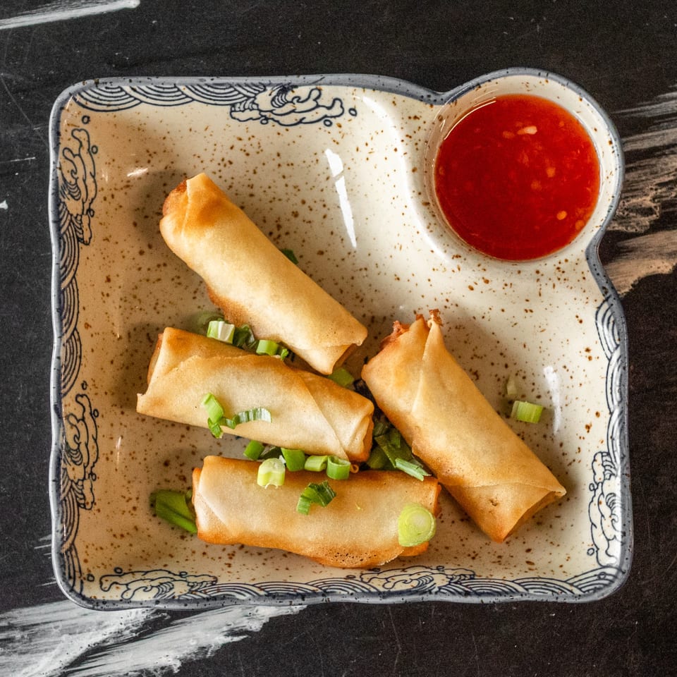 Four crispy spring rolls garnished with green onions, served with a side of red dipping sauce.