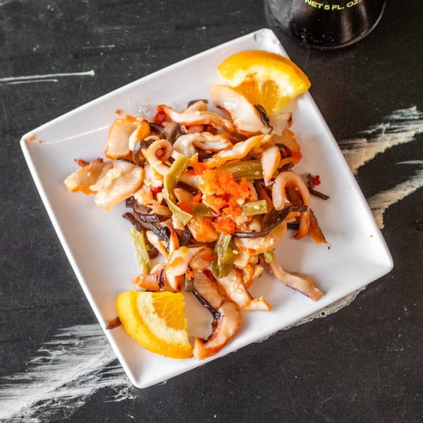 A white rectangular plate holds a stir-fry of seafood, vegetables, and orange tobiko, garnished with orange slices.