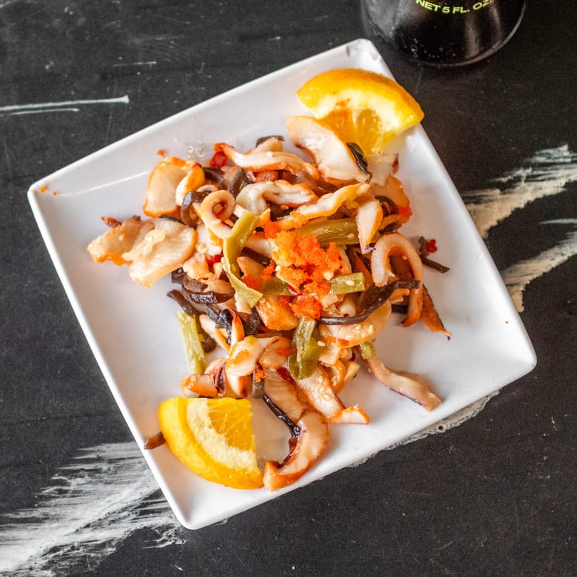 A white rectangular plate holds a stir-fry of seafood, vegetables, and orange tobiko, garnished with orange slices.