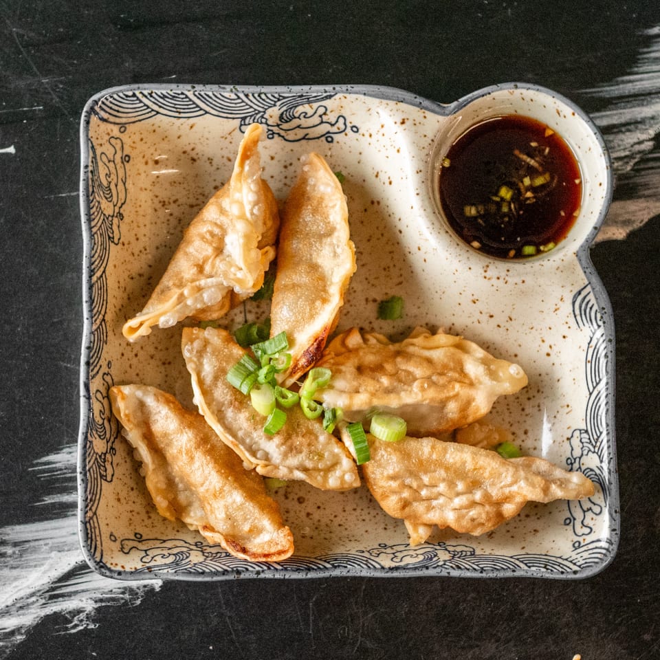 Golden-brown fried dumplings with green onions and a side of soy sauce on a decorative plate.